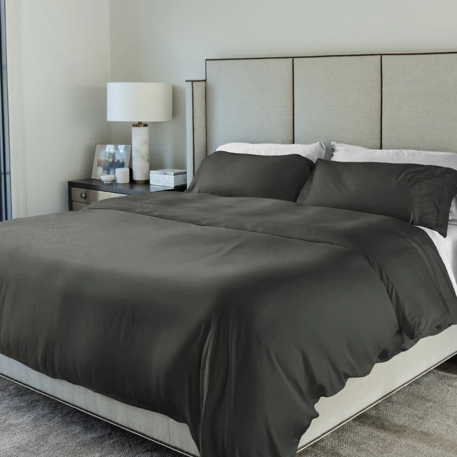 Gray bedding set on a bed in a bedroom with a lamp and nightstand.