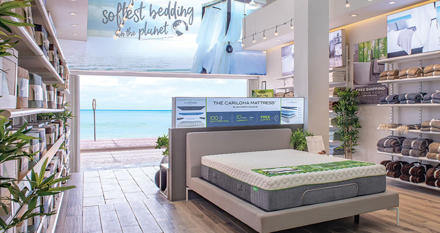 Cariloha retail store interior with a mattress display, folded bath towels on shelving, and an ocean view through the open doorway