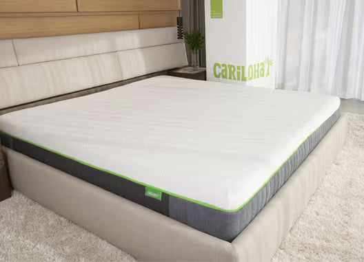 A Cariloha mattress on a matching upholstered bed frame in a modern bedroom setting with a light wood headboard, white curtains, and a white rug.