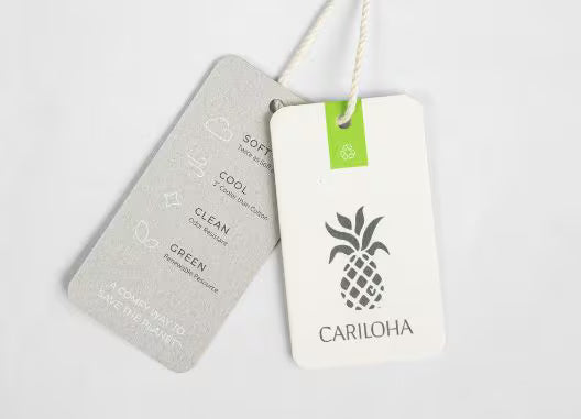 Two hanging product tags on a white cord against a light gray background. The front tag is white with a green square accent, a black pineapple graphic, and the brand name CARILOHA.
