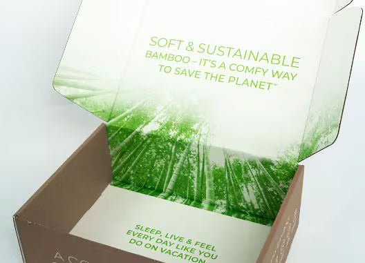 An open brown cardboard box with a printed bamboo forest design on the inside lid and bottom. Green text on the lid reads, 'SOFT & SUSTAINABLE BAMBOO - IT'S A COMFY WAY TO SAVE THE PLANET,' and text on the bottom reads, 'SLEEP, LIVE & FEEL EVERY DAY LIKE YOU DO ON VACATION.