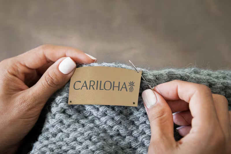 Close-up of a person's hands holding a gray knit garment and pinning a tan paper brand tag that reads CARILOHA.