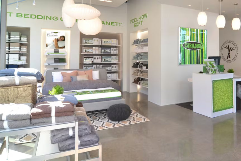 A bright bedding showroom interior with a bed, folded towels, shelves of bedding, hanging pendant lights, and a reception desk. 