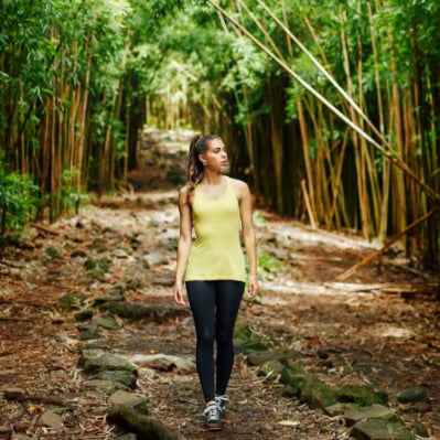A woman walking on a forest path surrounded by a dense bamboo grove, wearing a light yellow tank top and black leggings.