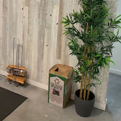 A tall artificial bamboo plant in a dark gray pot, placed next to a cardboard recycling bin against a light wood-paneled wall.