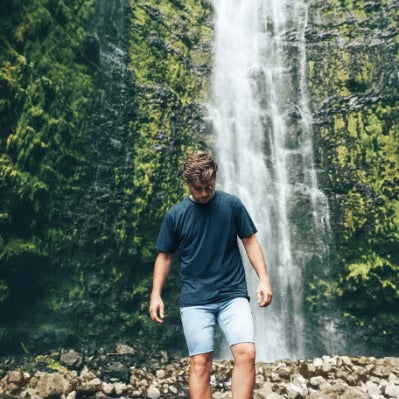 A man wearing a dark navy t-shirt and light blue shorts walks in front of a tall waterfall cascading down a mossy green cliffside.