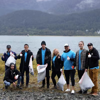 A group of nine volunteers posing on a rocky shoreline with mountains and water in the background, holding white trash bags. They are dressed in casual outdoor clothing, with several wearing matching bright blue shirts.