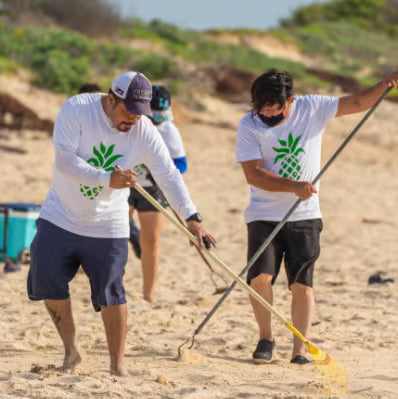 Two men on a sandy beach wearing matching white long-sleeve t-shirts with a green pineapple graphic, actively cleaning sand with brooms. One man is also wearing a dark cap and the other a black face mask.