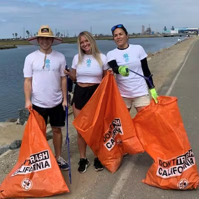 Three people standing on a waterfront path holding large orange trash bags printed with 'DON'T TRASH CALIFORNIA'