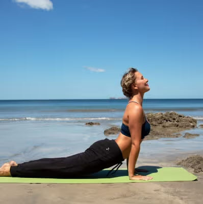 A woman in a navy sports bra and black yoga pants performs a cobra pose on a light green yoga mat at the beach, with the ocean and blue sky in the background.