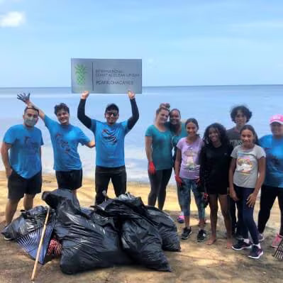 A group of volunteers stands on a beach after cleaning up trash, with several black trash bags in the foreground and a person holding a sign that reads 'International Day of Clean Ocean #carilohacares'.