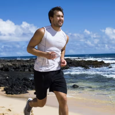 A man jogging on a sandy beach with the ocean and rocky shoreline in the background, wearing a white sleeveless athletic shirt and black shorts.