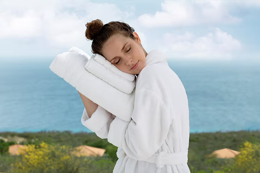 A woman in a white bathrobe hugging a folded white towel outdoors with the ocean and sky in the background.