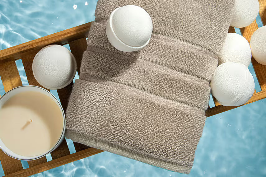 A beige terry towel draped over a wooden tray, surrounded by white bath bombs and a small candle, set against a bright blue water background.