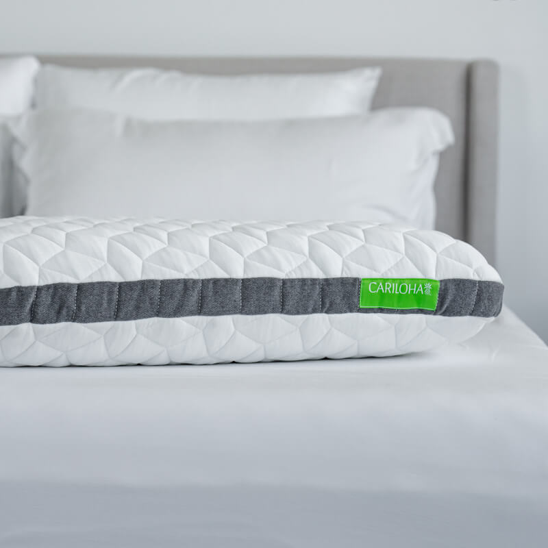 A white quilted memory foam pillow with a dark gray horizontal band and a green CARLOHA brand tag, resting on a bed with white pillows and a gray headboard.