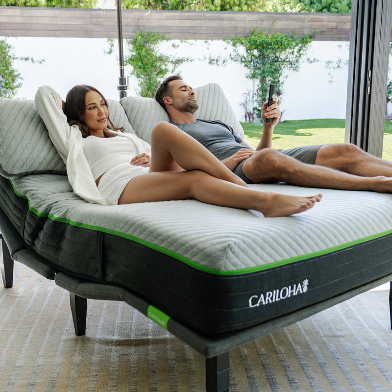 A man and a woman relax on a CARILOHA adjustable base and mattress in a modern indoor-outdoor living space with large windows and greenery outside.