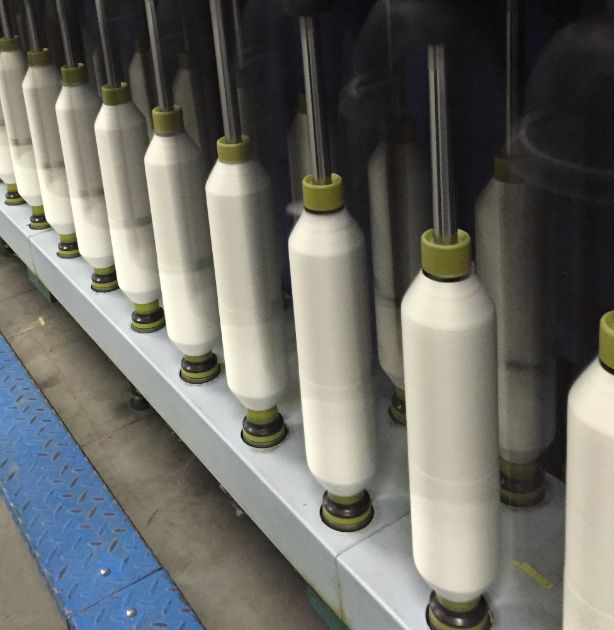 A close-up view of a textile spinning machine with multiple white spools of thread mounted on a long metal frame. The spools are wrapped in dark protective material near the top and bottom, with small olive-green caps.