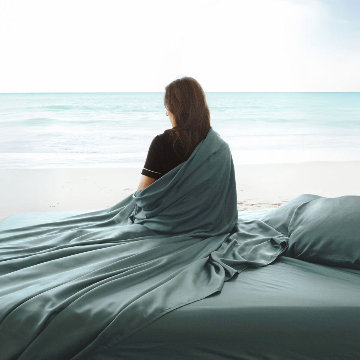 A woman sits on a bed wrapped in a teal bamboo sheets, facing a calm ocean view from a beach.