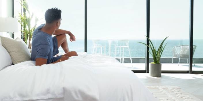 Man sitting on the edge of a white bamboo-bedded bed, gazing out floor-to-ceiling windows at the ocean